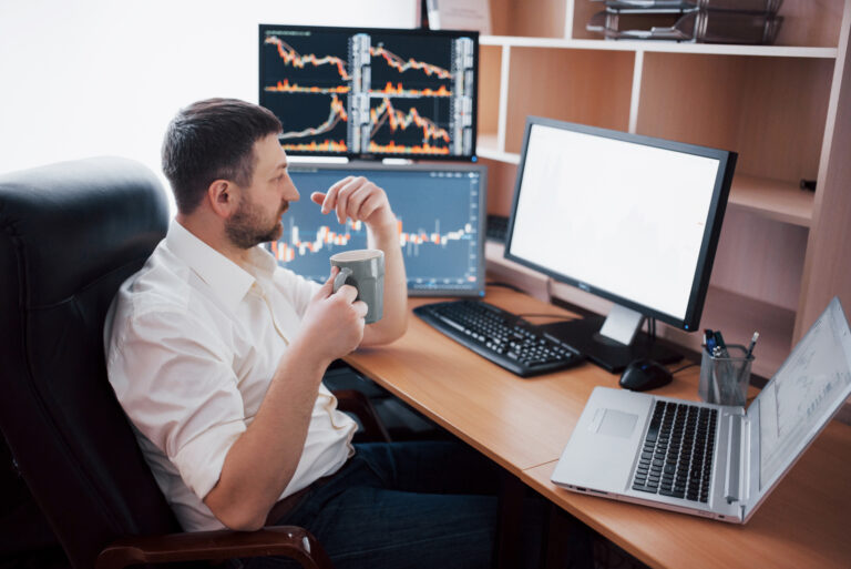 Young businessman is sitting in office at table, working on computer with many monitors,diagrams on monitor. Stock broker analyzes binary options charts